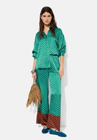 Zielona bluzka w groszki i pasujące spodnie z brązowymi akcentami. Modelka trzyma torebkę z frędzlami w odcieniu piaskowym, nosi niebieskie baleriny. Satynowy materiał o gładkiej teksturze.