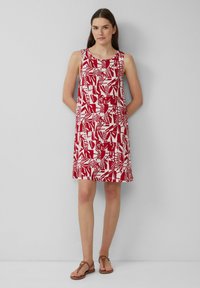 Czerwona i biała sukienka w wzory, bez rękawów, z okrągłym dekoltem, z abstrakcyjnymi kształtami i liśćmi. Noszona z brązowymi sandałami.