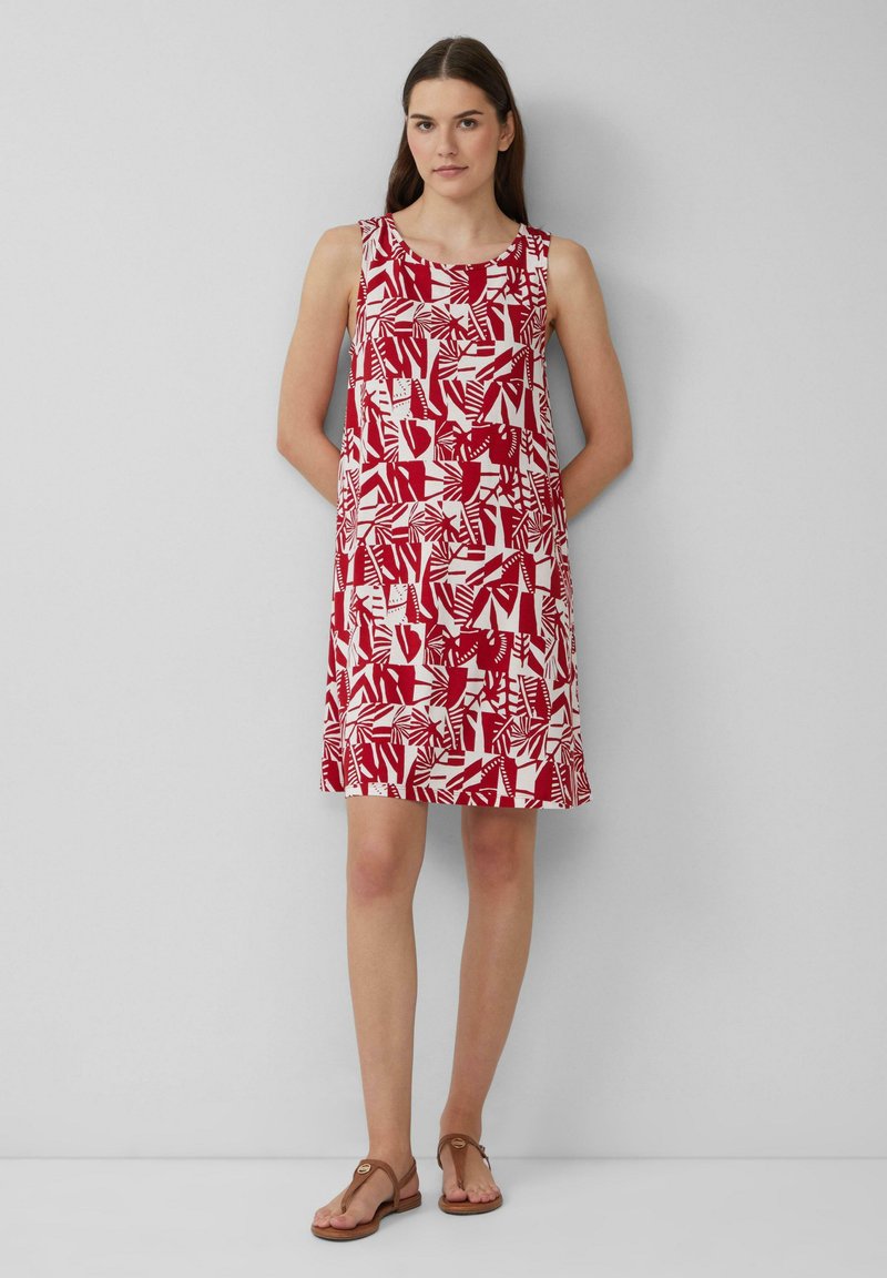 Czerwona i biała sukienka w wzory, bez rękawów, z okrągłym dekoltem, z abstrakcyjnymi kształtami i liśćmi. Noszona z brązowymi sandałami.