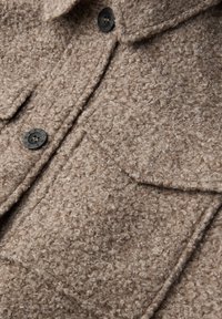 Hnědý kabát s texturou z měkkého, chlupatého materiálu, s černými knoflíky a dvěma kapsami na hrudi. Detailní prošívání podél švů.