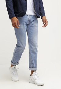 Jeans de denim azul claro con corte recto, doblados en el tobillo, combinados con una camiseta blanca y una chaqueta azul marino, complementados con zapatillas blancas.