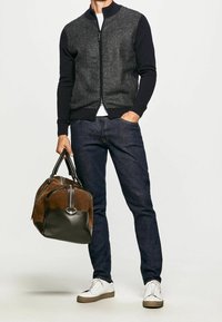 Herren Zip-Pullover mit strukturiertem grauen Körper, marineblauen Ärmeln, kombiniert mit dunklen Jeans. Hält eine braun-schwarze Leder-Reisetasche. Weiße Sneakers.