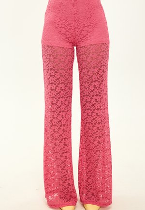 Pantalones de encaje floral rosa de pierna ancha con pantalones cortos ajustados visibles debajo, usados con zapatos de punta beige sobre un fondo claro.