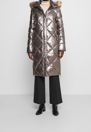 Long manteau matelassé métallisé avec capuche bordée de fourrure, porté sur un col roulé clair, pantalon noir à jambes larges et bottines noires.