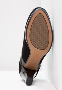 Chaussure à talon haut en cuir suédé noir et en cuir verni, avec une semelle marron texturée et le logo "Clarks", photographiée du côté de la semelle.
