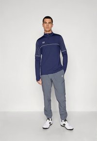 Tamsiai mėlynas sportinis megztinis su pusiau užtrauktuku ant apykaklės, baltais akcentais ir ilgomis rankovėmis, derinamas su pilkomis siaurėjančiomis kelnėmis ir baltais sportbačiais.