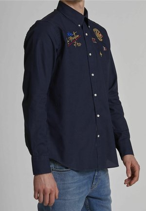 Homme portant une chemise bleu marine à boutons avec des écussons brodés sur la poitrine et un jean bleu clair, debout devant un fond gris uni.
