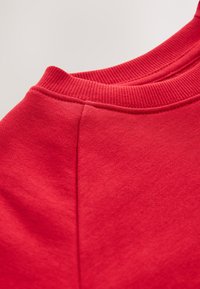 Punane dressipluus soonikkoelise kaelusega ja siledal pinnal. Omaduseks on õmbluste detail õlal, mis näitab kanga ühtlast paksust.