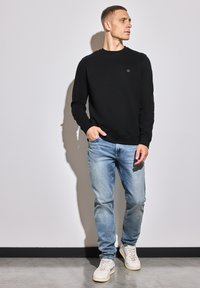 Schwarzer Rundhalsausschnitt-Pullover mit kleinem Logo auf der Brust, kombiniert mit hellblauen Jeans und weißen Sneakers.