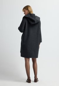 Tmavě šedý kabát s kapucí, volného střihu, délky po kolena, se dvěma postranními kapsami, vyrobený z texturované látky. Pod ním jsou vidět hnědé boty.