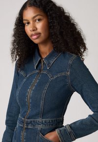 Giacca di denim blu scuro, con chiusura a zip, colletto a punta e cuciture definite. Maniche lunghe con estremità a polsino; include tasche laterali.