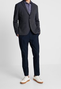 Blazer gris oscuro con dos bolsillos delanteros sobre una camisa con estampado floral. Combina con pantalones azul marino y zapatillas blancas con suelas marrones.