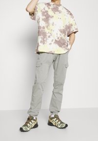 Batikt-shirt i gult, brunt och vitt; ljusgrå cargobyxor; gröna och vita träningsskor med gula detaljer och snören.