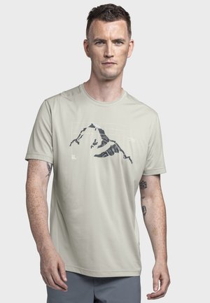 Mann trägt hellbeiges T-Shirt mit Berggrafik, kurzen Ärmeln und sichtbaren Tattoos auf den Unterarmen, steht vor schlichten Hintergrund.