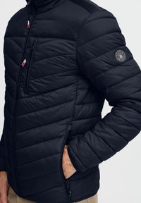 Svart pufferjakke med horisontalt quiltet mønster, glidelåslommer og et merke på ermet. Har kontrastfarger på skuldrene.