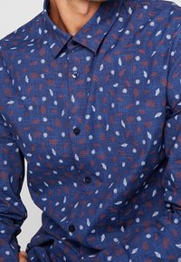 Koszula z długim rękawem w granatowym kolorze z teksturowanej tkaniny. Posiada rozsiane wzory liści w kolorze czerwonym i białym. W skład koszuli wchodzą czarne guziki i kołnierzyk.