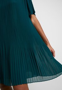 Tissu de robe plissée bleu sarcelle s'écoulant sur le bras et la cuisse, montrant la texture et le mouvement sur un fond uni.