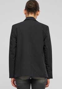 Czarna marynarka o strukturalnym kroju, z pojedynczym rozcięciem z tyłu i długimi rękawami. Materiał wydaje się gładki, bez widocznych wzorów czy detali metalowych.