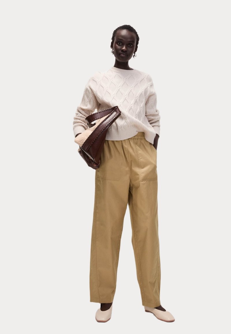 Pull en tricot crème avec un motif en losange, pantalon large beige et chaussures claires. Tenant un sac en cuir marron avec des détails en fourrure.