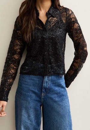 Femme portant une chemise noire en dentelle à manches longues avec boutons et détails en sequins, associée à un jean bleu taille haute.