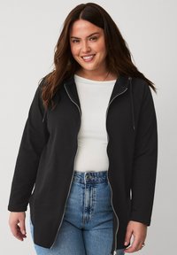 Sweat-shirt à zip noir en tissu lisse, avec capuche, fermeture éclair argentée et manches longues. Associé à un t-shirt blanc et un jean bleu.