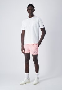 T-shirt bianca a maniche corte con taschino, abbinata a pantaloni corti rosa chiaro con logo. Il modello indossa sneakers bianche e calzini sportivi.