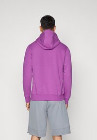 Sweatshirt roxa com um grande capuz, mangas longas e bainha canelada. Usado com calções cinzentos, exibindo um estilo atlético casual.