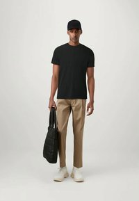 Muž v černé kšiltovce, černém tričku, béžových kalhotách a bílých teniskách, držící černou prošívanou tašku proti jednobarevnému světlému pozadí.