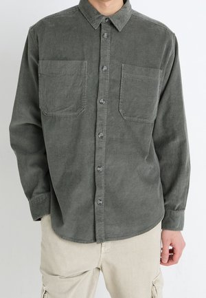 Osoba ubrana w ciemnoszarą koszulę z długim rękawem wykonaną z sztruksu, z dwoma kieszeniami na piersi oraz jasnobeżowe spodnie cargo.