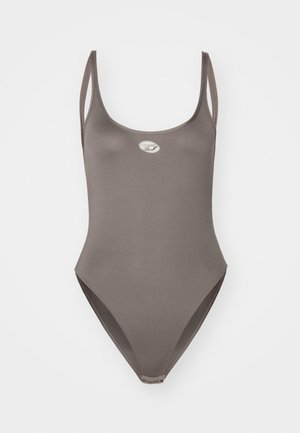 Maillot de bain en tissu marron clair, conçu avec des fines bretelles, des jambes hautes et un petit logo métallique à l'avant. Texture lisse.