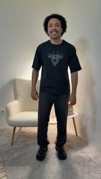 Jeune homme aux cheveux bouclés souriant, portant un t-shirt noir avec motif, un pantalon noir et des bottes, debout dans une pièce avec un fauteuil et une table d'appoint.