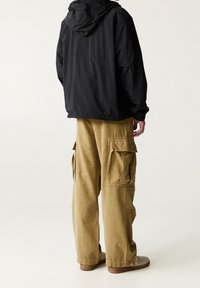 Giacca nera con cappuccio sopra pantaloni cargo marroni chiari con molte tasche laterali; vestibilità ampia; tessuto morbido; calzature marroni con punta arrotondata.