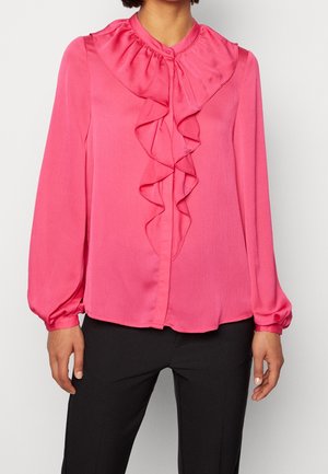 Vaaleanpunainen pusero, jossa on korkea röyhelökaulus, pitkät hihat ja napillinen etuosa; valmistettu sileästä, kevyestä kankaasta.