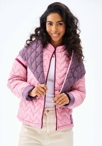 Žena sa usmieva, má na sebe ružovo-fialovú prešívanú bundu cez biele tričko a krémové nohavice, drží okraje bundy oboma rukami.