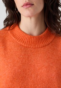 Oransje ullgenser med ribbestrikket rund hals, med en myk, teksturert overflate. Stoffet ser varmt og komfortabelt ut.