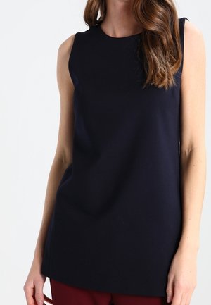 Haut noir sans manches à encolure ronde porté par une personne aux longs cheveux bruns ondulés, associé à un pantalon rouge foncé.