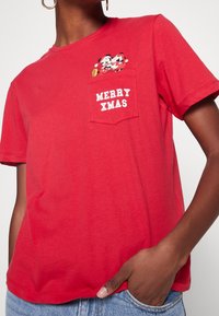 Camiseta de algodón roja con cuello redondo, que presenta un bolsillo en el pecho y el texto "MERRY XMAS" en blanco, junto a un diseño gráfico divertido.