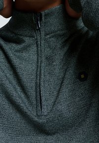 Gros plan sur une personne portant un pull zippé texturé gris avec un col partiellement fermé et un petit logo hexagonal sur la poitrine.