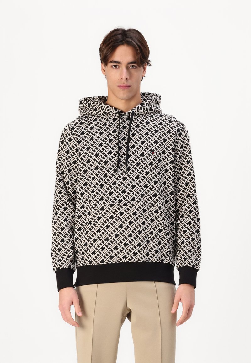 Felpa a motivo bianco e nero con cappuccio regolabile, polsini a coste e tasca a marsupio, indossata sopra pantaloni chiari.