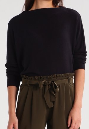 Vrouw draagt een zwart, lang mouw topje in een hooggesneden olijfgroene broek, vastgeknoopt met een stoffen riem in de taille.