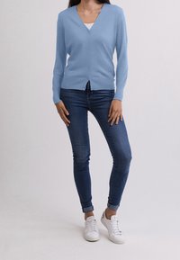 Person som bär en ljusblå knappad cardigan, vit skjorta under, mörkblå tajta jeans och vita sneakers, som står mot en enfärgad bakgrund.