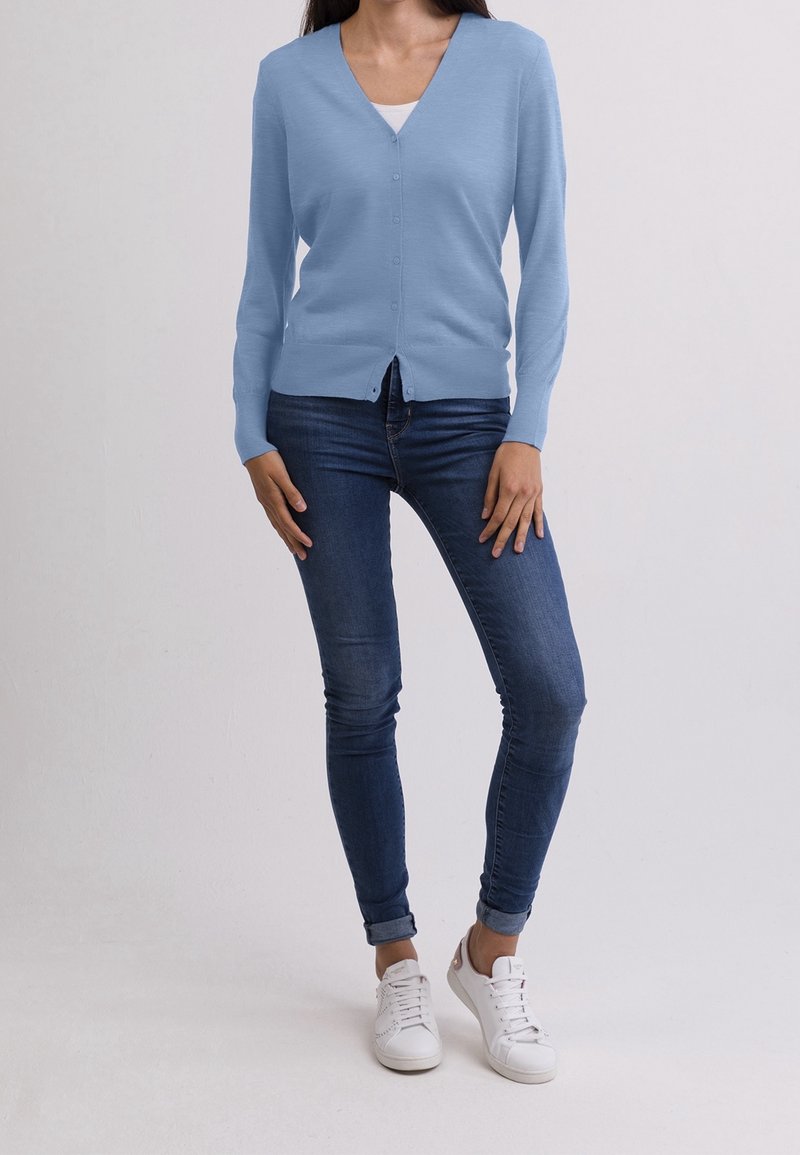 Person som bär en ljusblå knappad cardigan, vit skjorta under, mörkblå tajta jeans och vita sneakers, som står mot en enfärgad bakgrund.