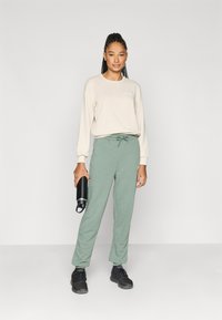 Lys beige langermet skjorte, grønne joggebukser med snøring i midjen, svarte joggesko, holder en svart vannflaske. Glatte stoffteksturer.