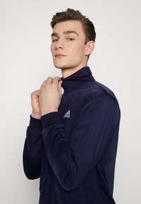 Chaqueta de zip azul marino con un alto cuello, puños acanalados y un logotipo de Adidas en el lado izquierdo del pecho. Fabricada con un tejido liso y elástico.