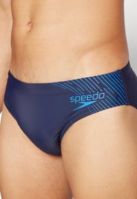 Férias de natação em azul marinho, feitas de material elástico, apresentam um logo da Speedo e um padrão dinâmico de riscas azuis na cintura.