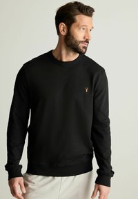 Sudadera negra de manga larga, con cuello redondo y puños de canalé. Presenta un pequeño emblema de ciervo en bronce en la parte izquierda del pecho.