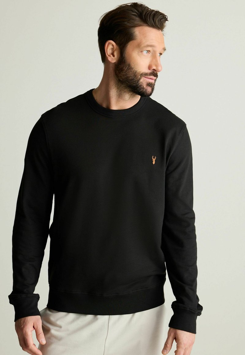 Sudadera negra de manga larga, con cuello redondo y puños de canalé. Presenta un pequeño emblema de ciervo en bronce en la parte izquierda del pecho.