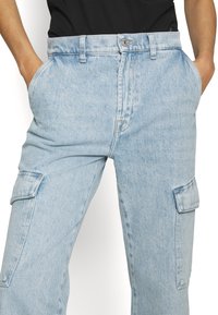 Pantalones cargo de mezclilla azul claro con un corte relajado, que cuentan con bolsillos laterales, pasadores para cinturón y un cierre de botón. Tejido texturizado con acentos desgastados.