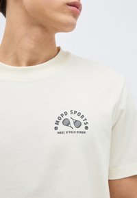 Κοντινό πλάνο ατόμου που φοράει εκρού μπλουζάκι με το κείμενο «MOPD SPORTS» και δύο σταυρωτά ρακέτες τένις πάνω από το λογότυπο «MARC O’POLO DENIM».