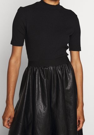 Vrouw draagt een aansluitend zwart geribd kortemouwtopje gecombineerd met een zwarte neplederen rok met hoge taille en een geplooide taille.